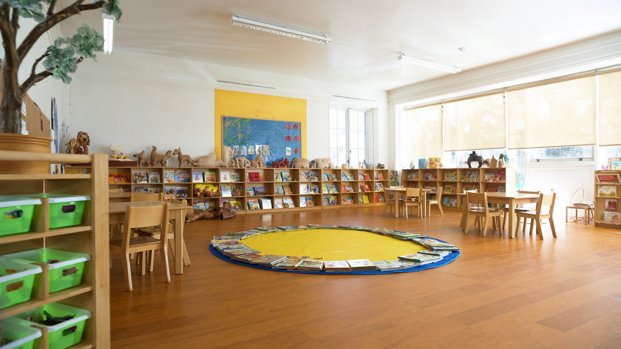 Bright, sunlit Bramblewood classroom with wood floors, low bookshelves full of books and toys, and a soft round play rug