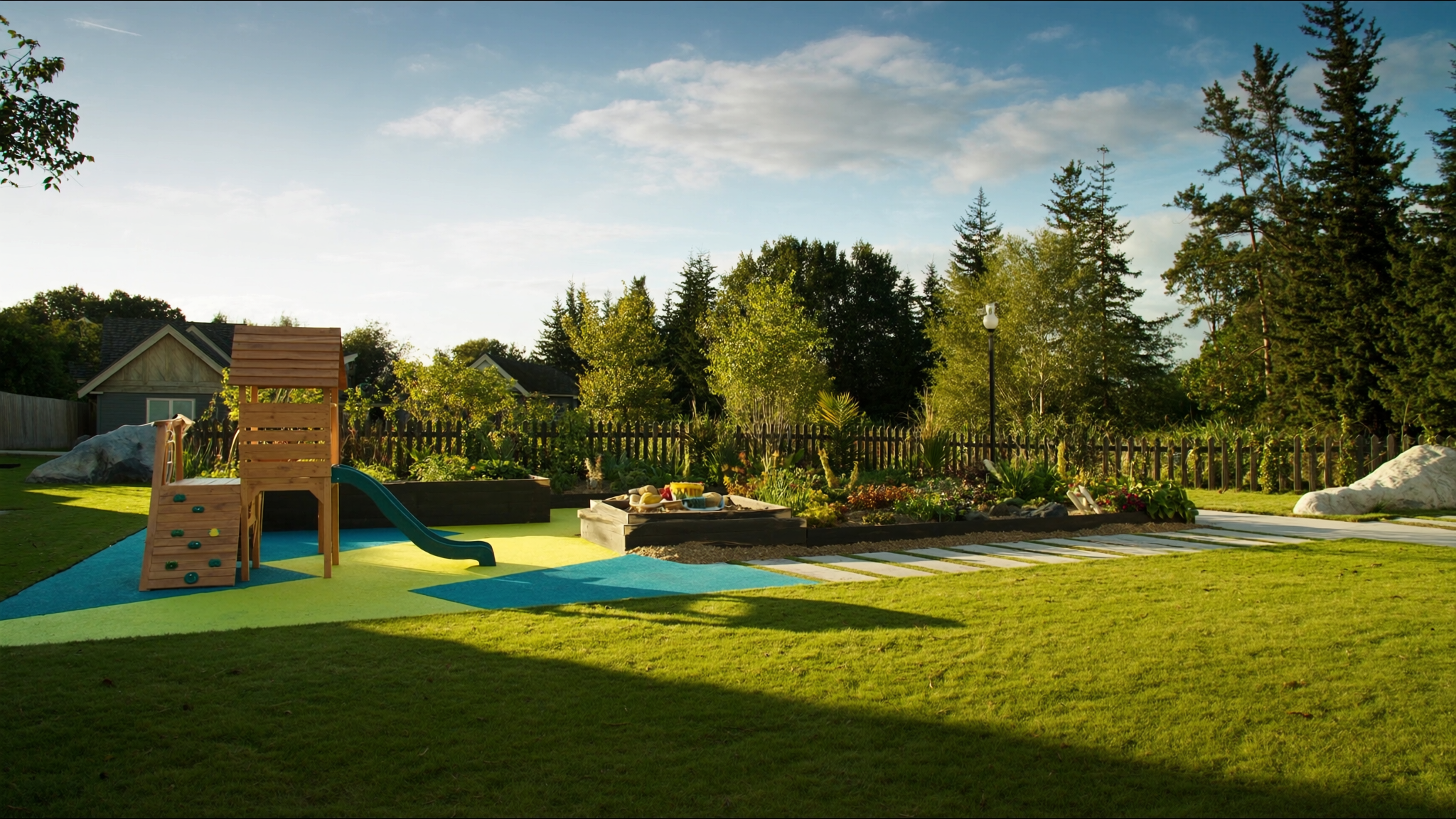 Fenced backyard playground with a wooden climbing structure, slide, garden beds, and green lawn in evening sun
